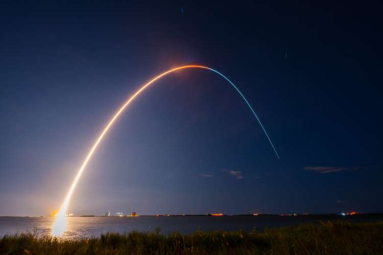 The image captures a nighttime rocket launch. The scene features a long-exposure photograph that shows the bright, curved trail of the rocket as it ascends into the dark sky. The trail starts from the brightly lit launch pad on the horizon near the left side of the image and arcs gracefully across the sky, fading towards the right. The sky is mostly clear, with a few scattered clouds and faint stars visible. In the foreground, there is a grassy area leading to a body of water that reflects the light.