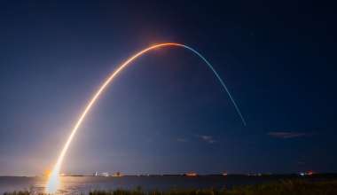 The image captures a nighttime rocket launch. The scene features a long-exposure photograph that shows the bright, curved trail of the rocket as it ascends into the dark sky. The trail starts from the brightly lit launch pad on the horizon near the left side of the image and arcs gracefully across the sky, fading towards the right. The sky is mostly clear, with a few scattered clouds and faint stars visible. In the foreground, there is a grassy area leading to a body of water that reflects the light.