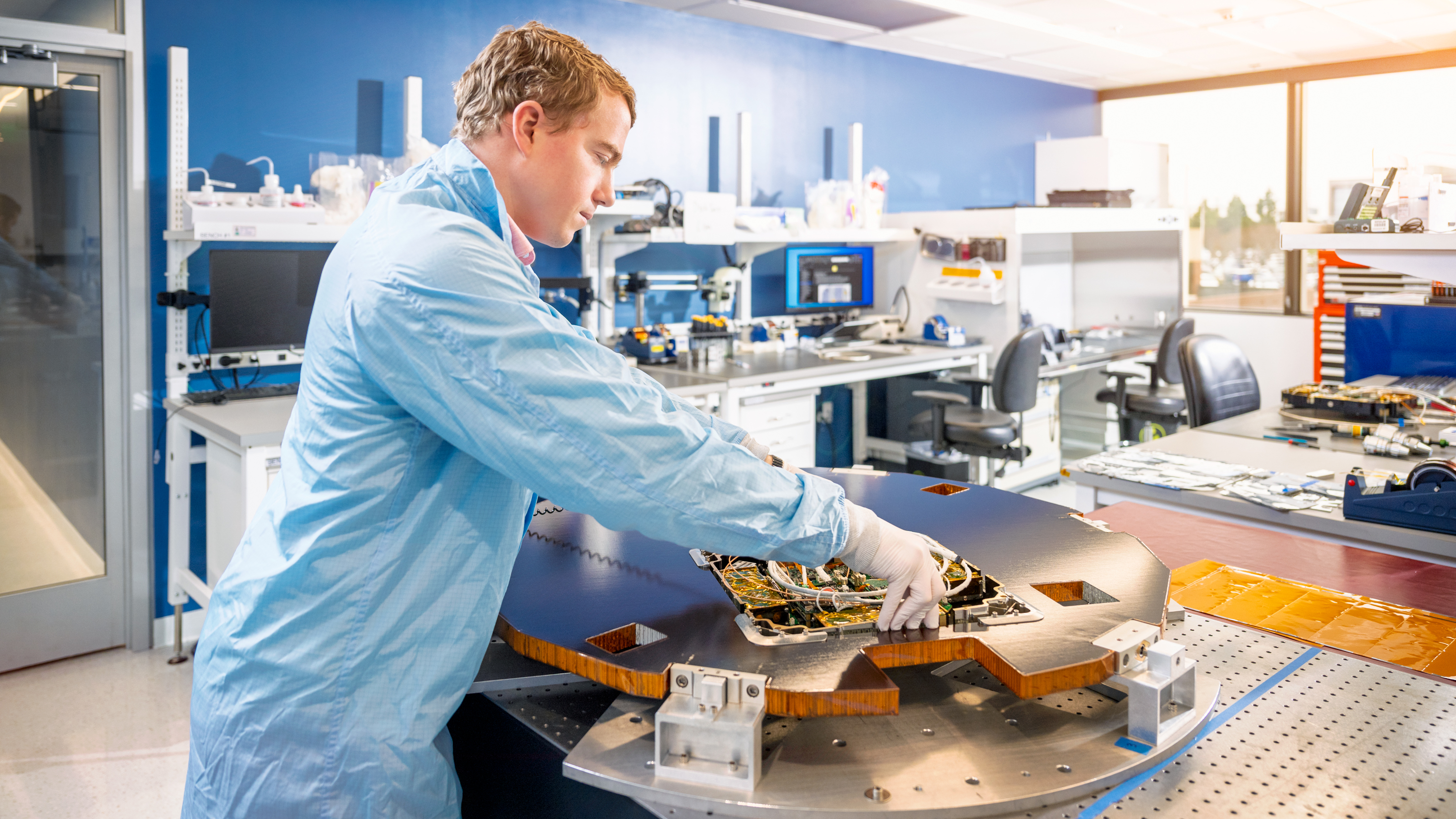 A person in a blue lab coat and white gloves is working in a high-tech laboratory or cleanroom. They are handling a large flat disk-shaped satellite on a specialized table. The background features organized workstations with monitors, tools, and other electronic devices, indicating a professional and controlled environment. The room is brightly lit with natural light streaming through large windows.