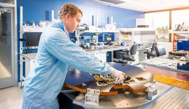 A person in a blue lab coat and white gloves is working in a high-tech laboratory or cleanroom. They are handling a large flat disk-shaped satellite on a specialized table. The background features organized workstations with monitors, tools, and other electronic devices, indicating a professional and controlled environment. The room is brightly lit with natural light streaming through large windows.